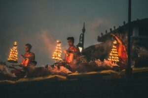 Three men in red robes performing a ritual with flaming tiered lamps at night
